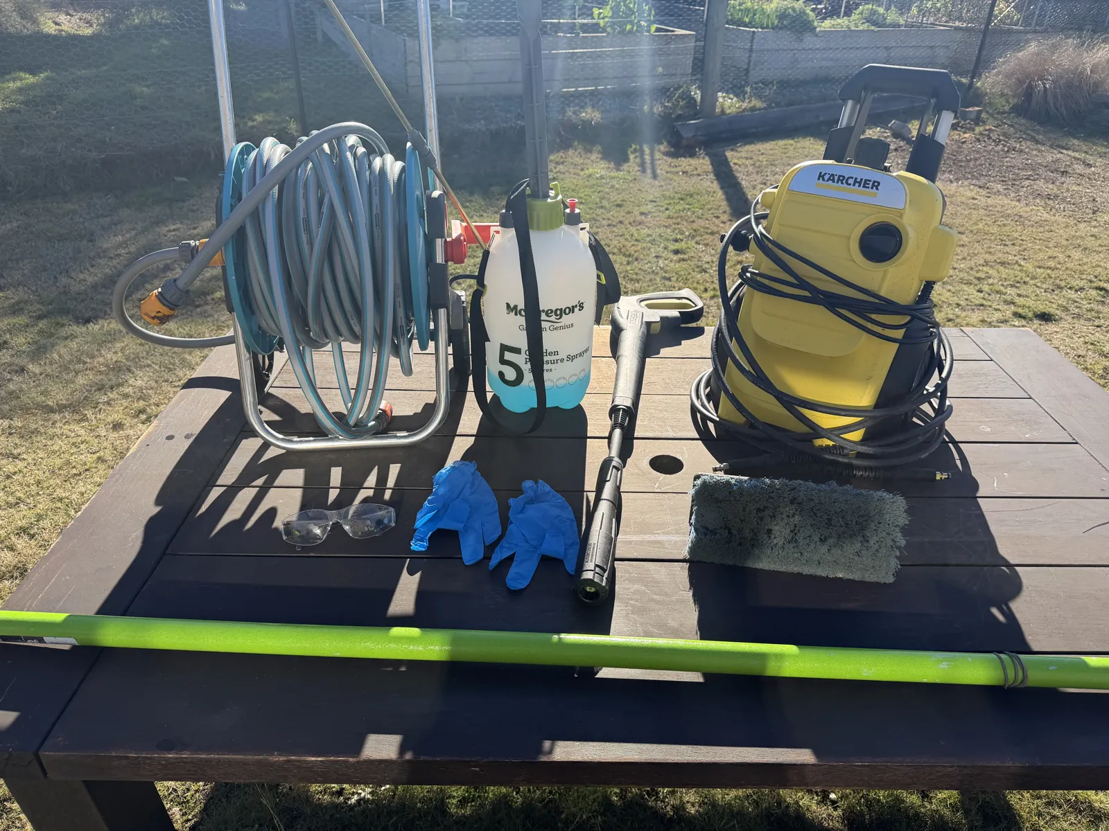Cedar washing kit laid out on a table: garden hose reel, 5 litre garden pressure sprayer, Karcher pressure washer with lance, soft-bristle brush head, safety glasses and gloves